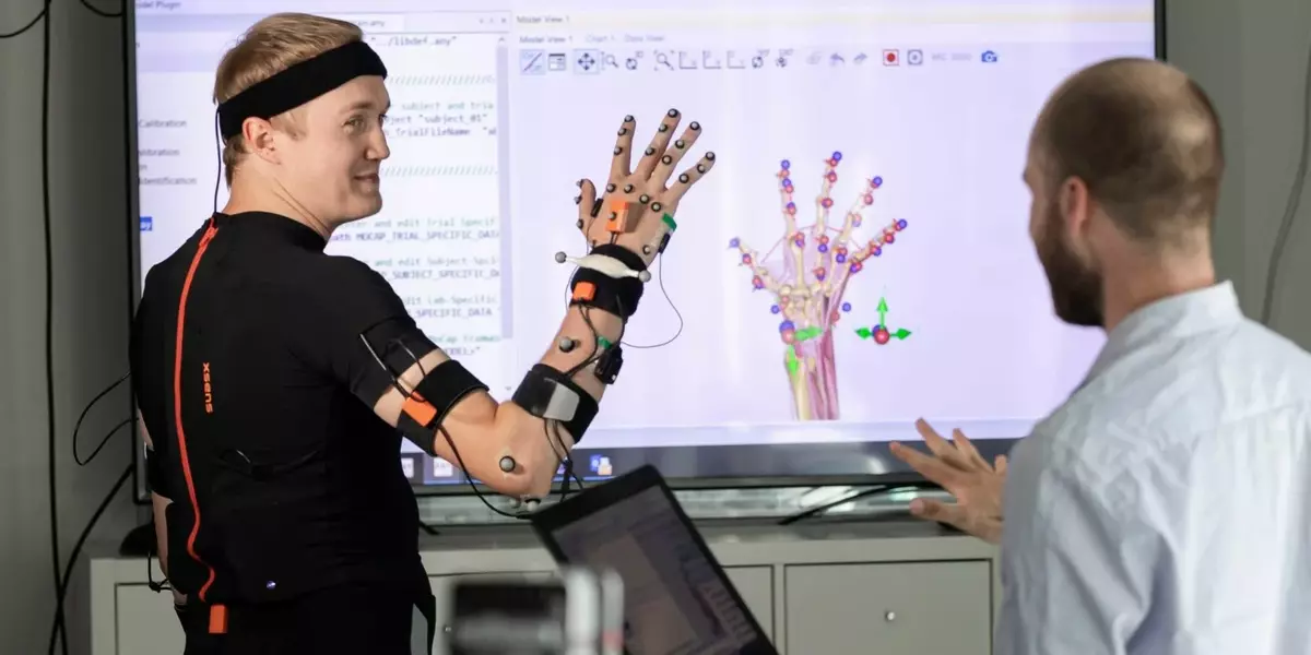 Ein Studierender hält seien verkabelte Hand in der Luft. Im Hintergrund wird auf einem Bildschirm die Biomechanik der Hand angezeigt.
